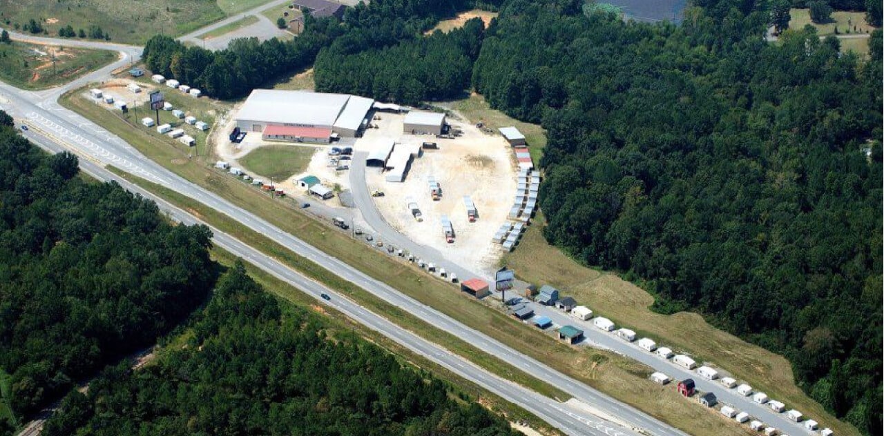 Aerial view of a large lot with several buildings, vehicles, and trailers, surrounded by dense forest. A highway runs alongside the lot, with cars traveling in both directions. The setting appears to be a rural area.