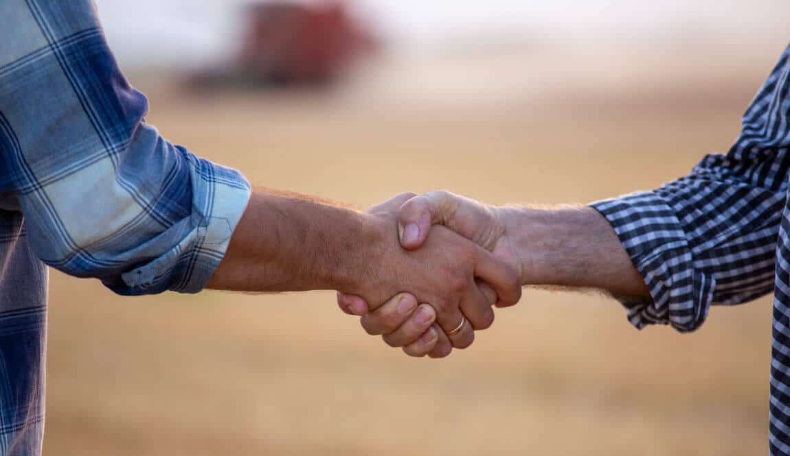 Two people shaking hands outdoors, both wearing plaid shirts. One shirt is blue and the other is black and white. The background is blurred, with earthy tones, suggesting a rural or natural setting.