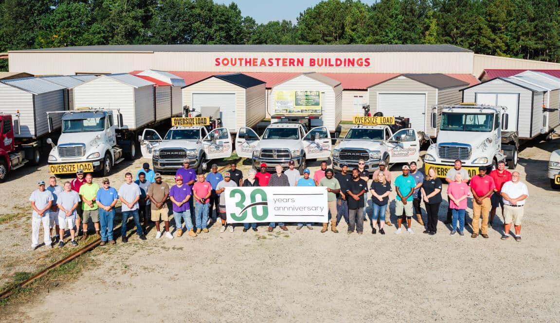 A group of people stand in front of trucks and a building with Southeastern Buildings signage. They hold a banner celebrating the 30th anniversary. The background features trees and a clear sky.