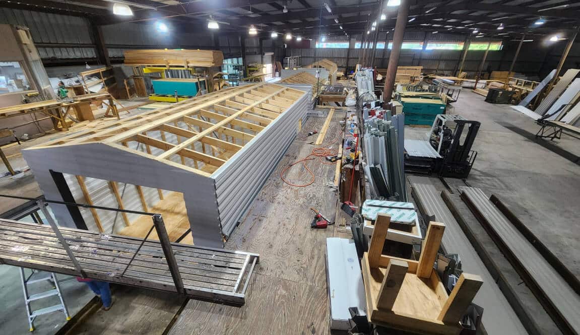 A large factory interior with workers assembling prefabricated houses. Wooden frames are visible, along with various tools and materials. The environment is spacious, with high ceilings and overhead lighting.