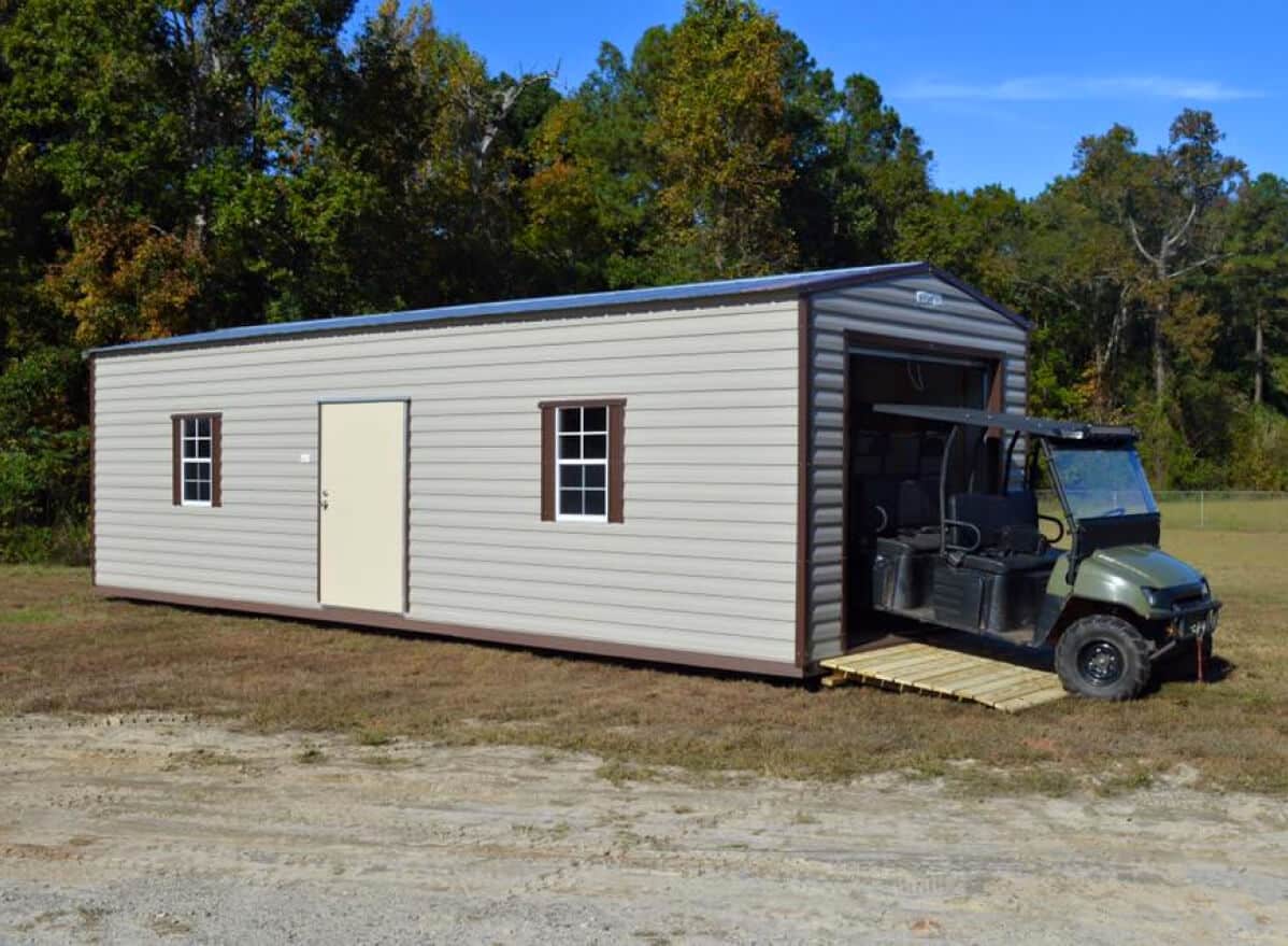 A long, light-colored storage shed with two windows and a door sits on grassy ground. A small utility vehicle partially enters the shed via a wooden ramp. Trees with green and autumn foliage are visible in the background.