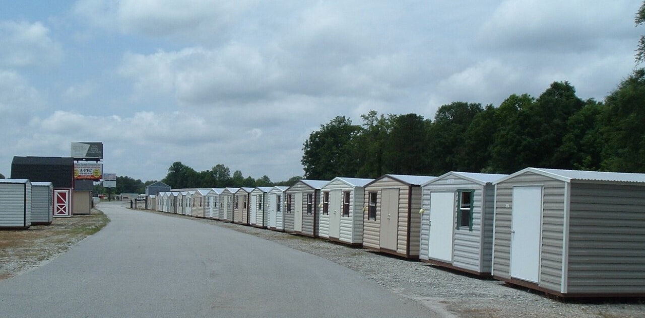 A row of small sheds lined up along a paved road on a cloudy day. The sheds vary in color, and there are trees in the background. A few signs are visible near the sheds.