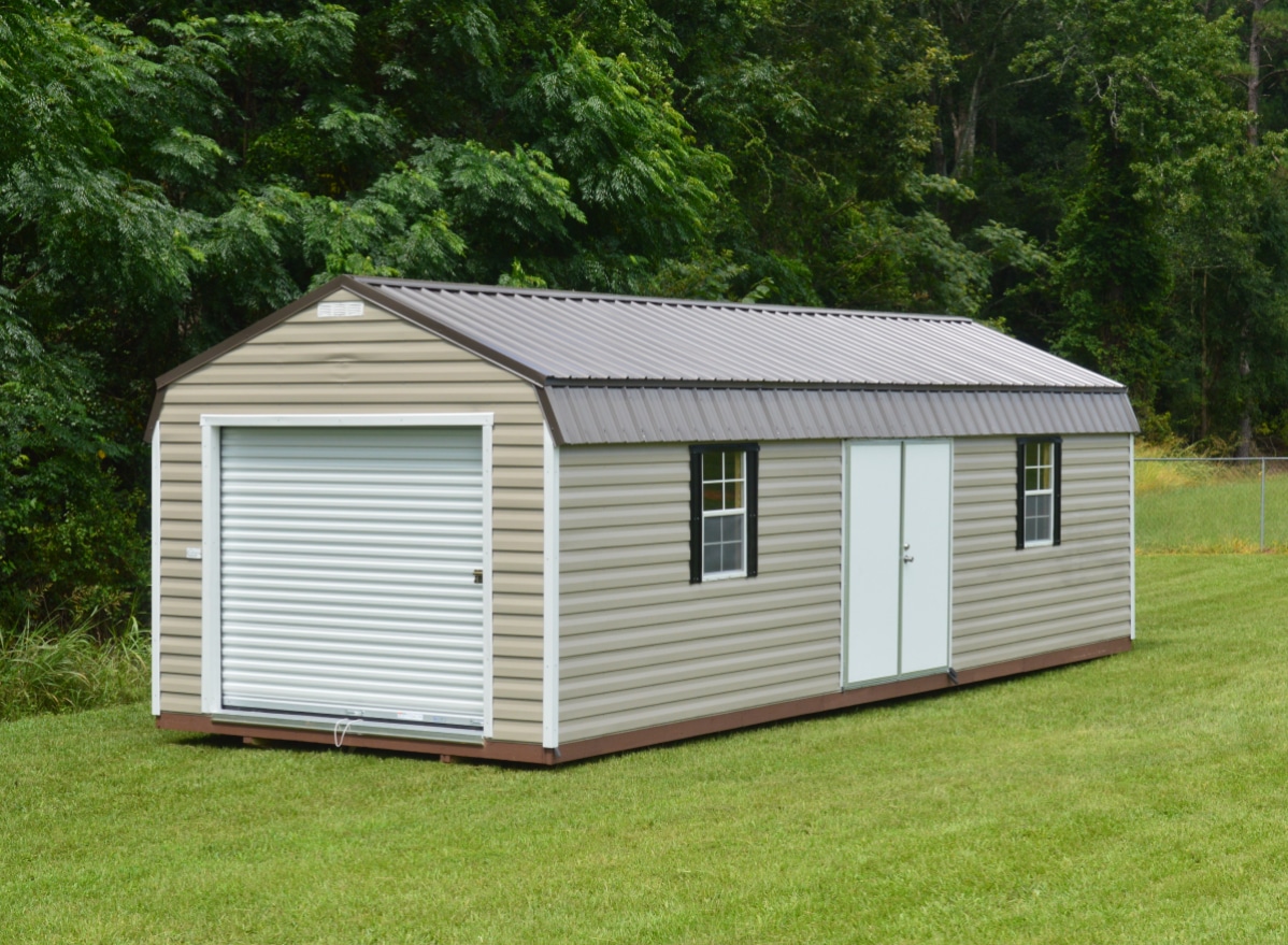 A small light beige shed with a metal roof and a porch stands on a grassy field. It has a wooden deck and supports, a glass-paneled door, and trees in the background under a partly cloudy sky.