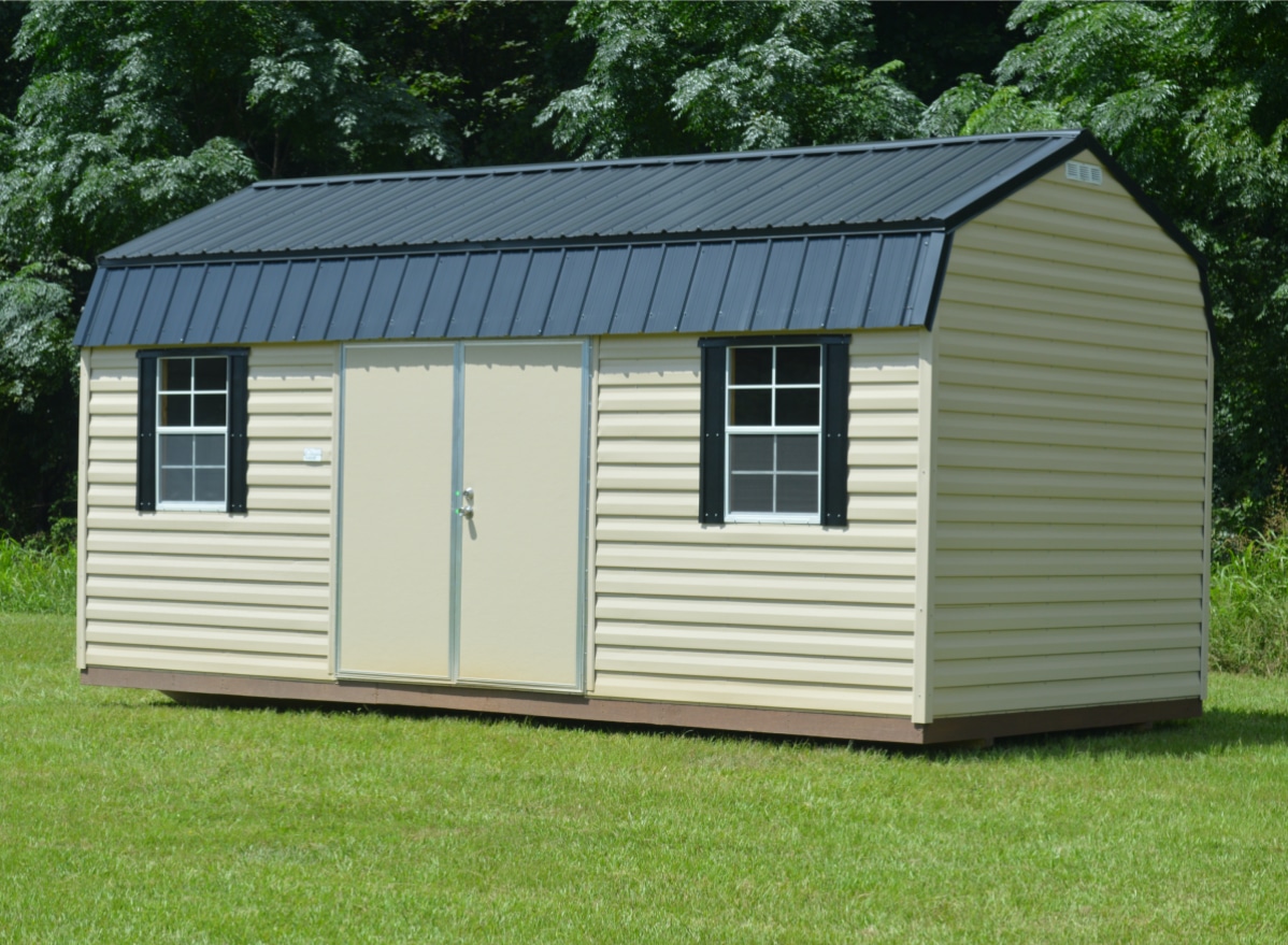 Alt text: A beige shed with a black metal roof and two windows with shutters. It stands on a grassy area, surrounded by trees. The shed has a central door and appears new and well-maintained.