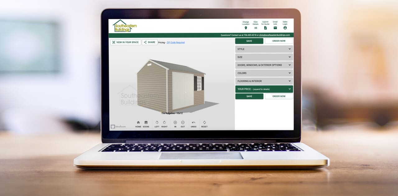 A laptop displaying a shed design tool on its screen, showing a 3D model of a gray shed. The interface includes options for style, doors, colors, and accessories, with buttons to save and order now. The laptop sits on a wooden surface.