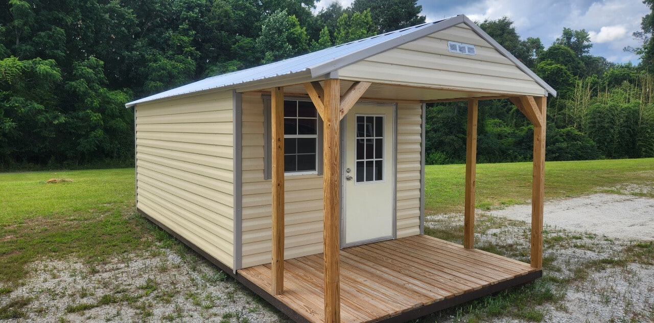 A small, beige shed with a metal roof and wooden porch set on a grassy area. It has a white door with a window and is surrounded by trees and greenery under a partly cloudy sky.