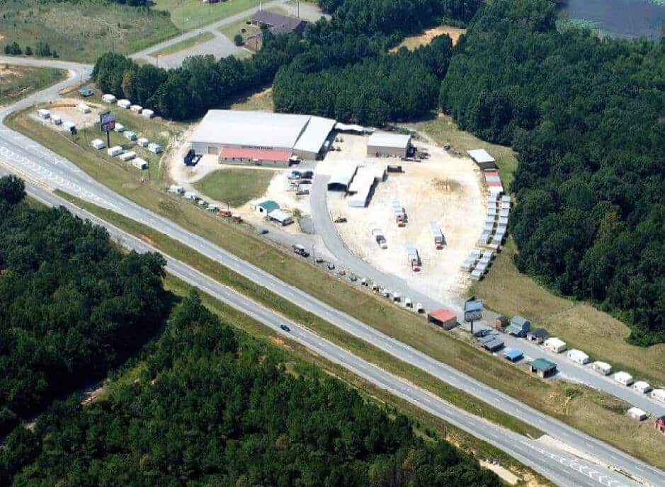 Aerial view of a large facility with multiple white buildings and a dirt parking lot, surrounded by dense green forest. A two-lane highway runs parallel to the site, with several vehicles visible near the buildings.