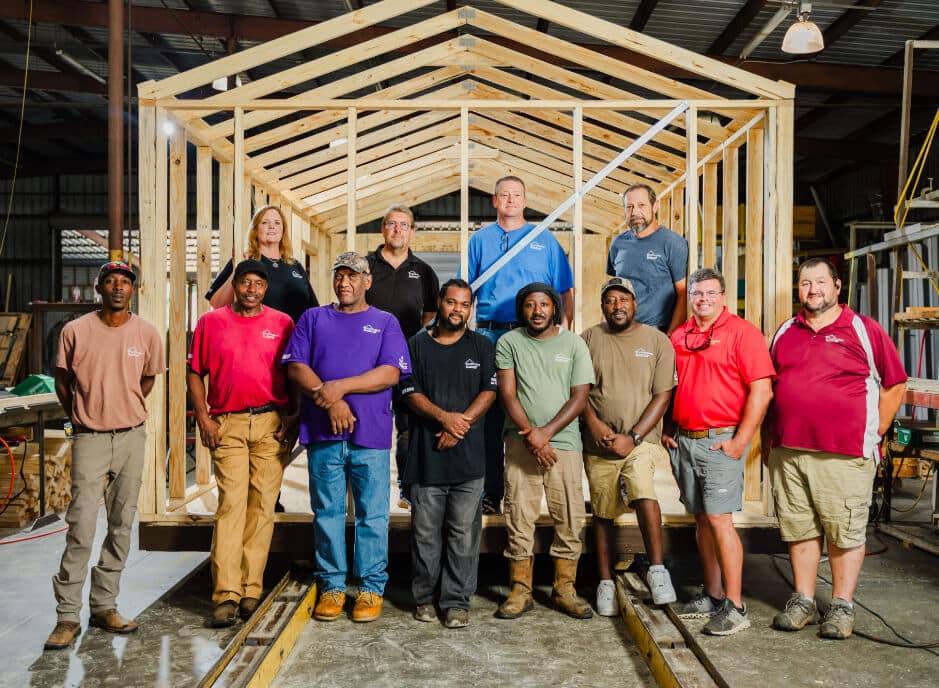 A diverse group of 12 people stands in front of a wooden frame of a tiny house inside a workshop. They are wearing casual clothing in various colors, and the background shows construction materials and equipment.