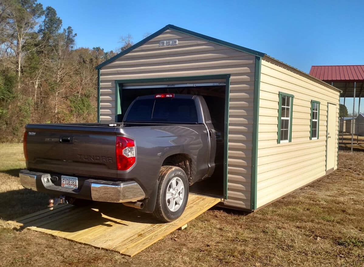 A gray pickup truck is partially backed out of a narrow beige and green portable garage or shed on a grassy field. The truck's rear is visible, and the garage has small windows on the side.