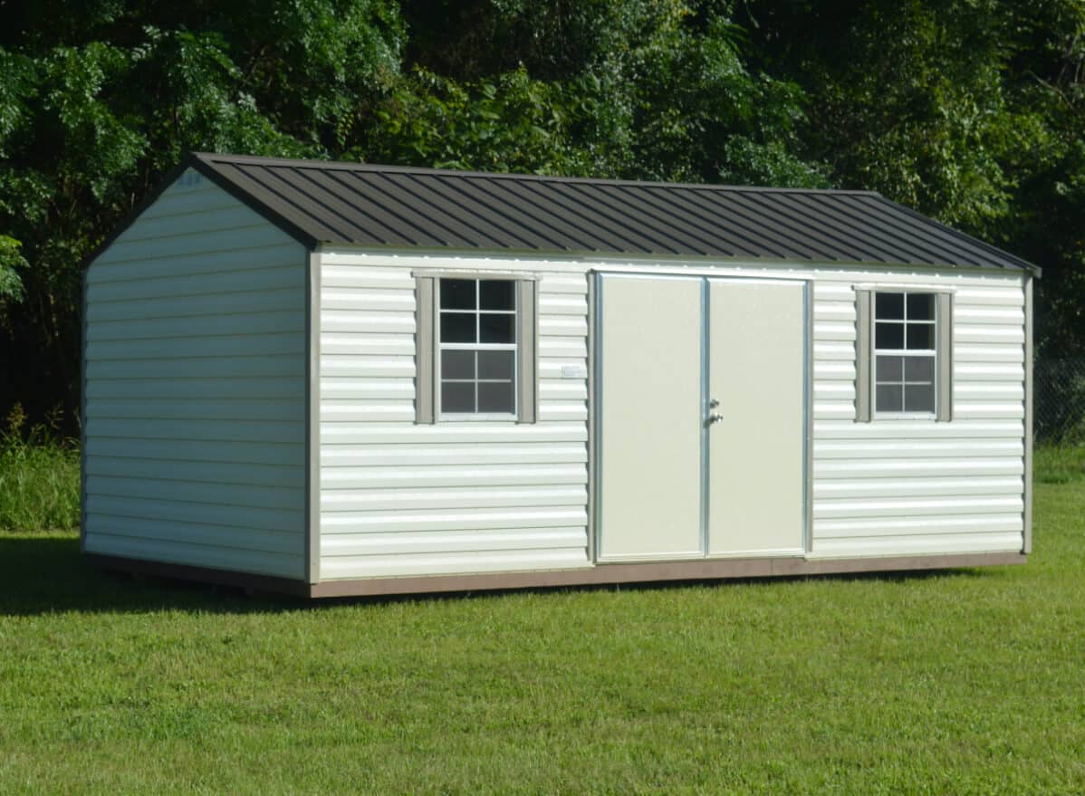 A white and gray outdoor storage shed with a metal roof, two windows, and a double door. It is situated on a grassy lawn with dense green foliage in the background.