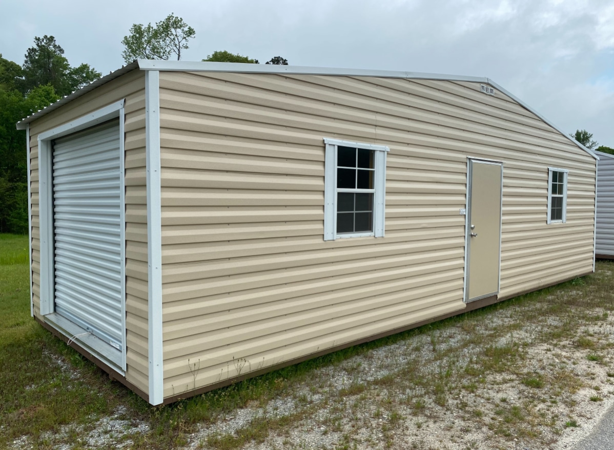 A beige metal storage shed with a white roll-up garage door on the left, a window in the middle, and a single door on the right. It is situated on grassy terrain with some gravel patches. Trees and a cloudy sky are in the background.