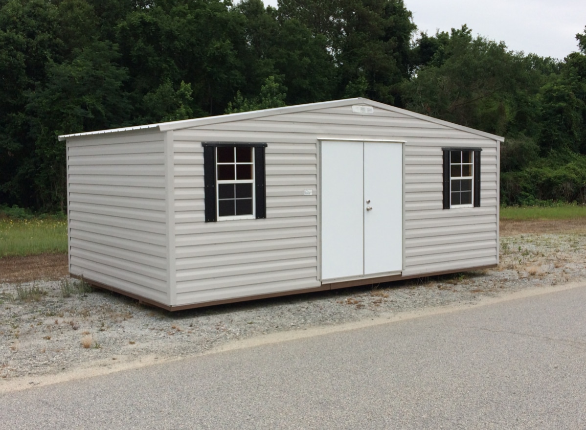 A gray, single-story metal shed with white framed windows and a double door sits on a gravel surface. It's surrounded by greenery and trees in the background.