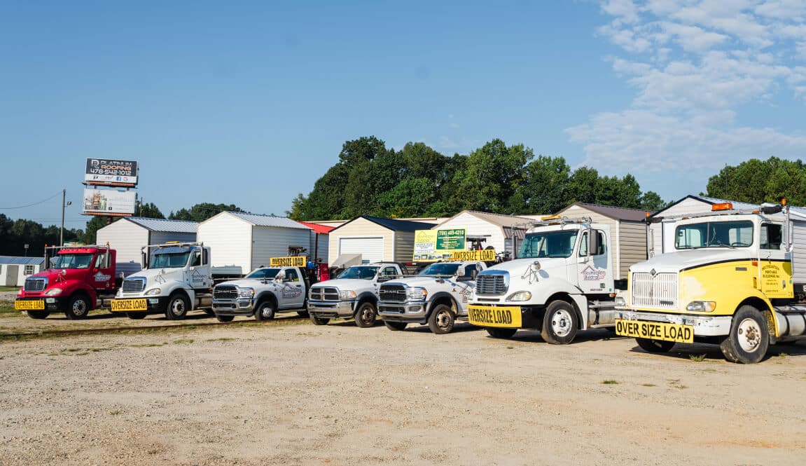 A lineup of seven trucks, including pickup and heavy-duty models, parked on a gravel lot. Each truck displays an OVERSIZE LOAD banner. Storage buildings and trees are visible in the background under a blue sky.