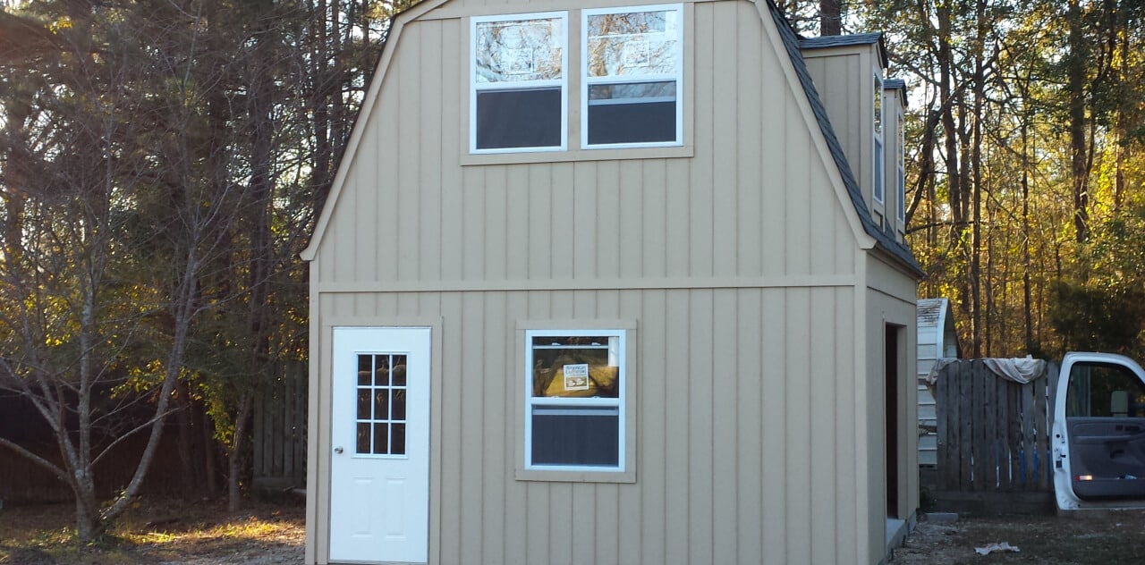 A light brown, two-story barn-style building with three windows and a white door stands in a wooded area. An open van door is visible on the right. The sun filters through trees, casting shadows on the building.