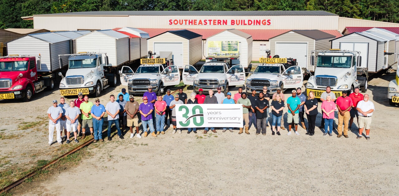 A group of people stands in front of several trucks and portable buildings, holding a banner that reads 30 years anniversary. The setting is outdoors, in front of a building marked Southeastern Buildings.