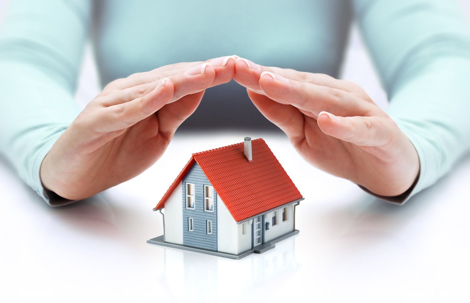 A pair of hands forms a protective gesture over a small model house with a red roof. The background is softly blurred, emphasizing the house and hands.